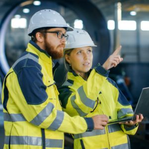 Portrait of Male and Female Industrial Engineers in Hard Hats Discuss New Project while Using Laptop. They Wear Safety Jackets.They Work at the Heavy Industry Manufacturing Factory.